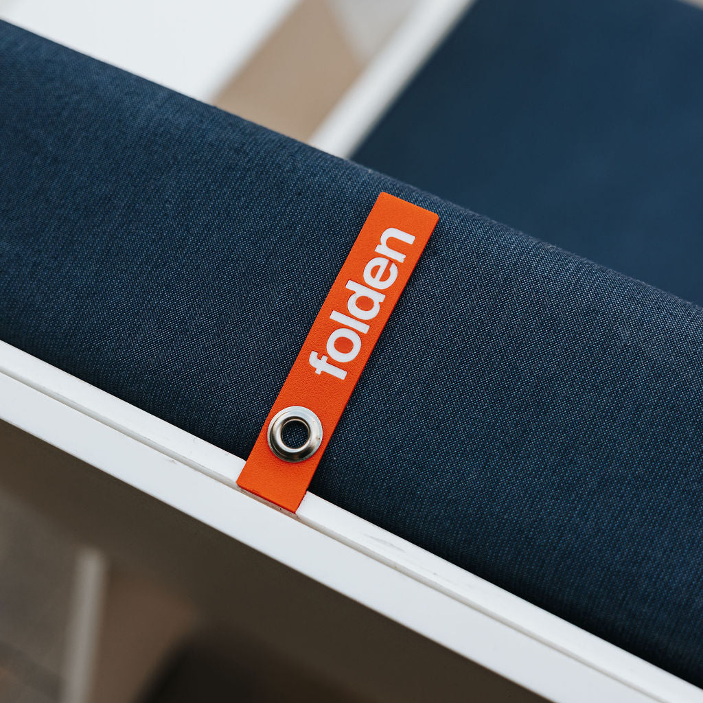 Orange FOLDEN pull strap with a metal eyelet against a dark blue back cushion and white chair frame.