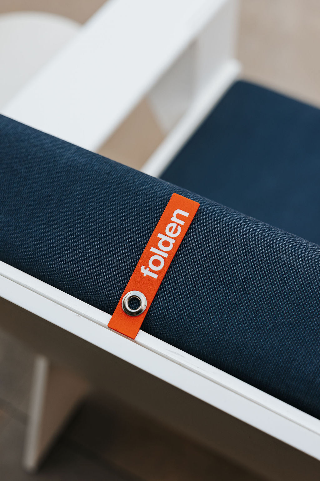 Orange FOLDEN pull strap with a metal eyelet against a dark blue back cushion and white chair frame.