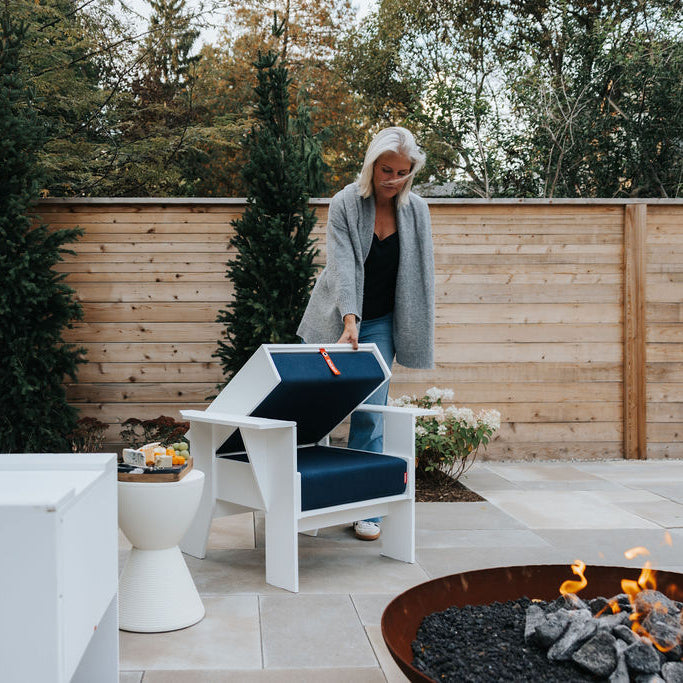 White FOLDEN Club Chair partway through folding, with a woman holding the backrest beside a patio fire pit.