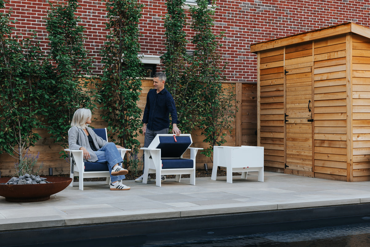 White FOLDEN Club Chairs on a patio, with a woman seated, a man closing one chair and another chair sealed for cushion storage.
